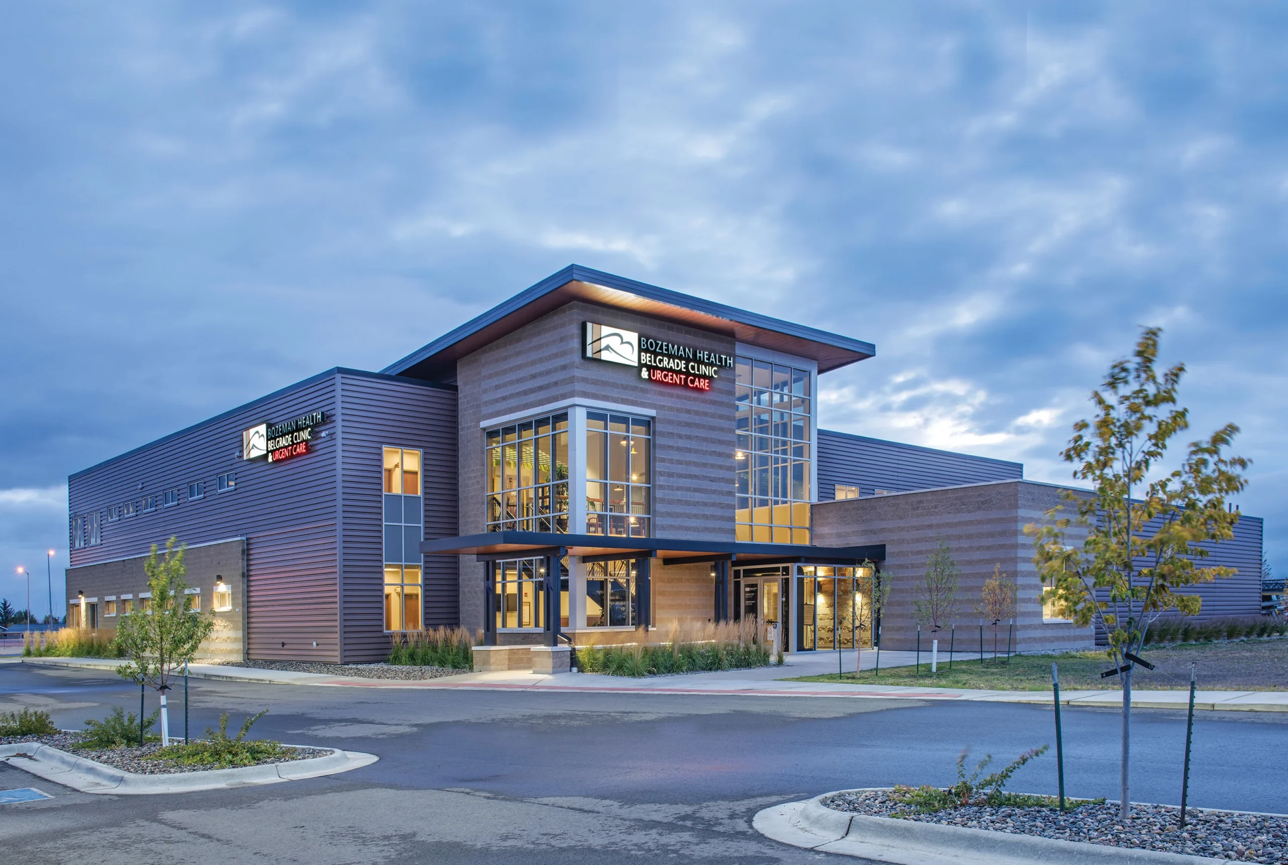 A modern exterior view of Bozeman Health Belgrade Clinic at dusk, showcasing its sleek architectural design with expansive glass windows, metal paneling, and warm interior lighting.