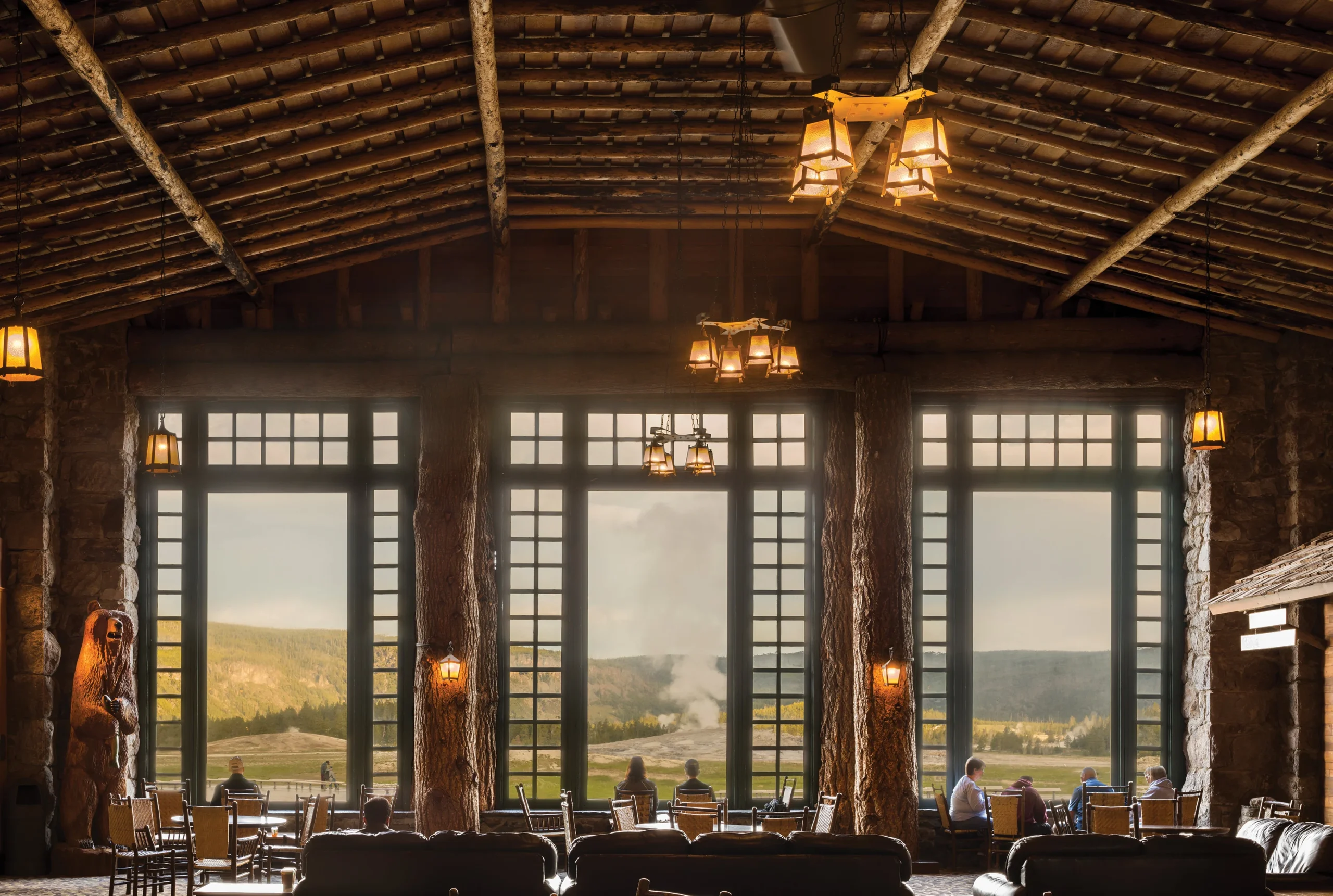 Majestic great room at Old Faithful Lodge showcasing rustic log architecture, towering floor-to-ceiling windows, and handcrafted lantern-style chandeliers.