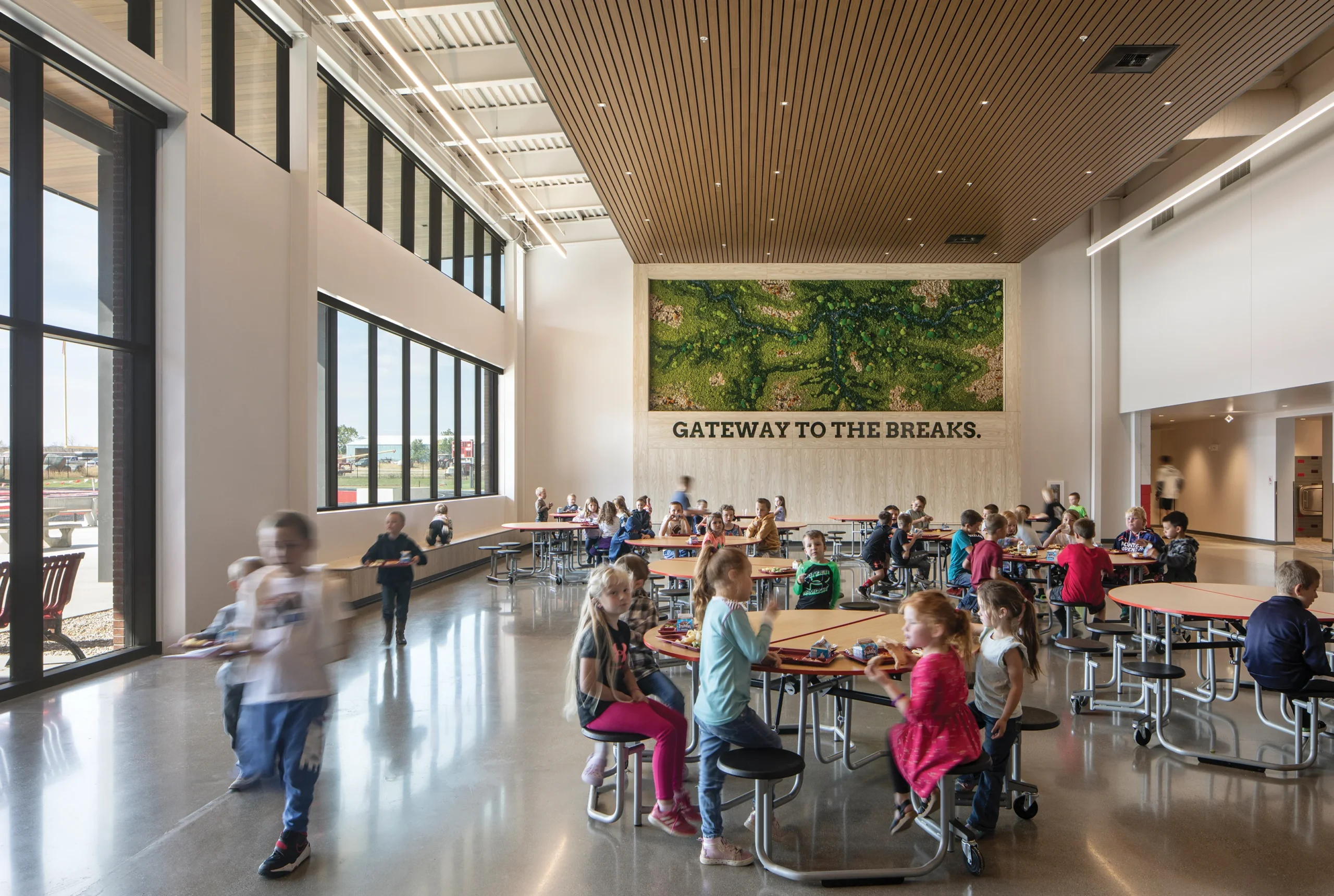 Bright, spacious cafeteria at Winifred Public Schools featuring natural light, modern furnishings, and a striking green art installation reading 'Gateway to the Breaks.' Students gather and socialize in a welcoming environment designed to inspire community.