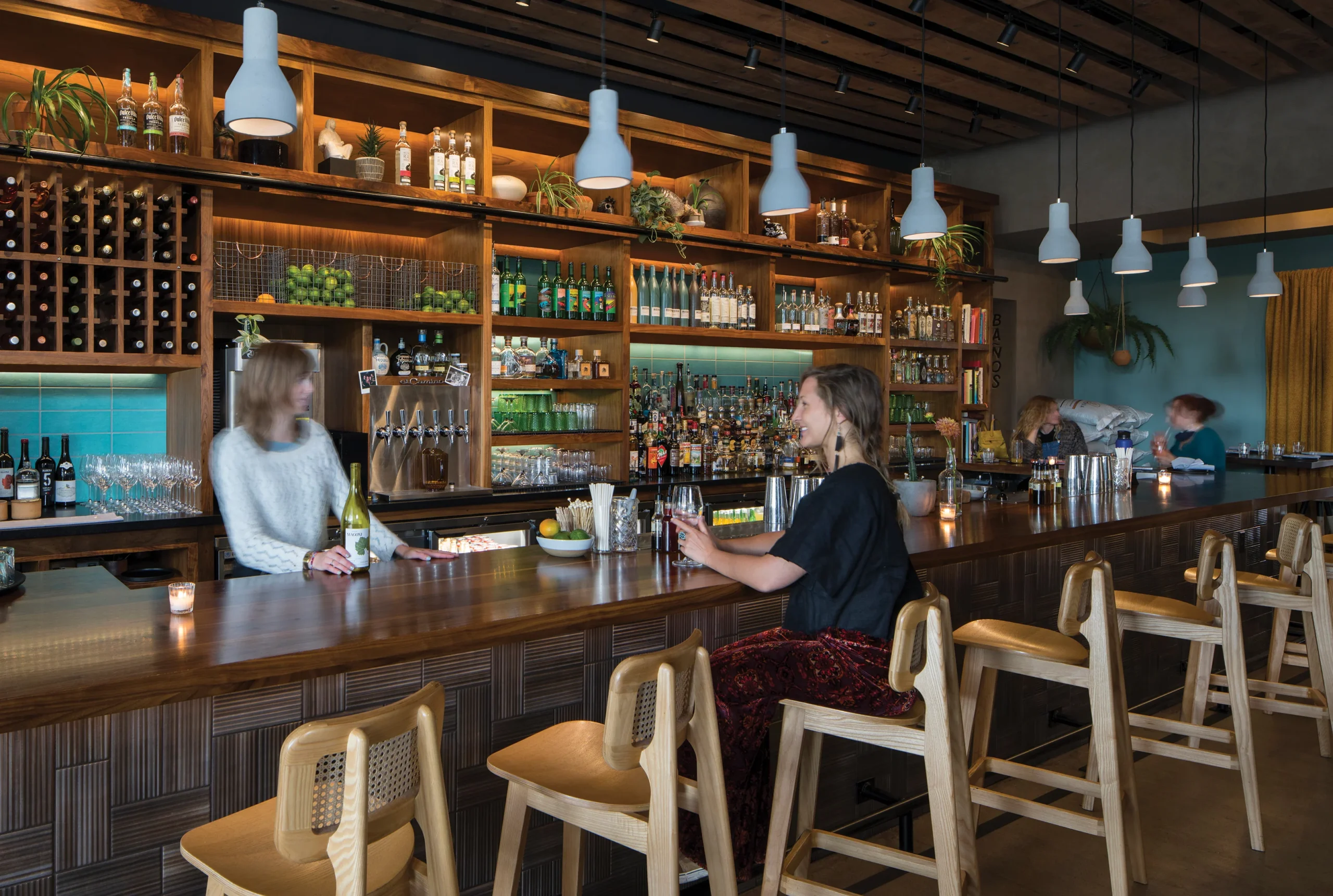 Modern Mexican-style bar interior at The Camino in Missoula, featuring a reclaimed wood bar, open shelving with spirits and greenery, and warm pendant lighting that enhances the vibrant, social ambiance.