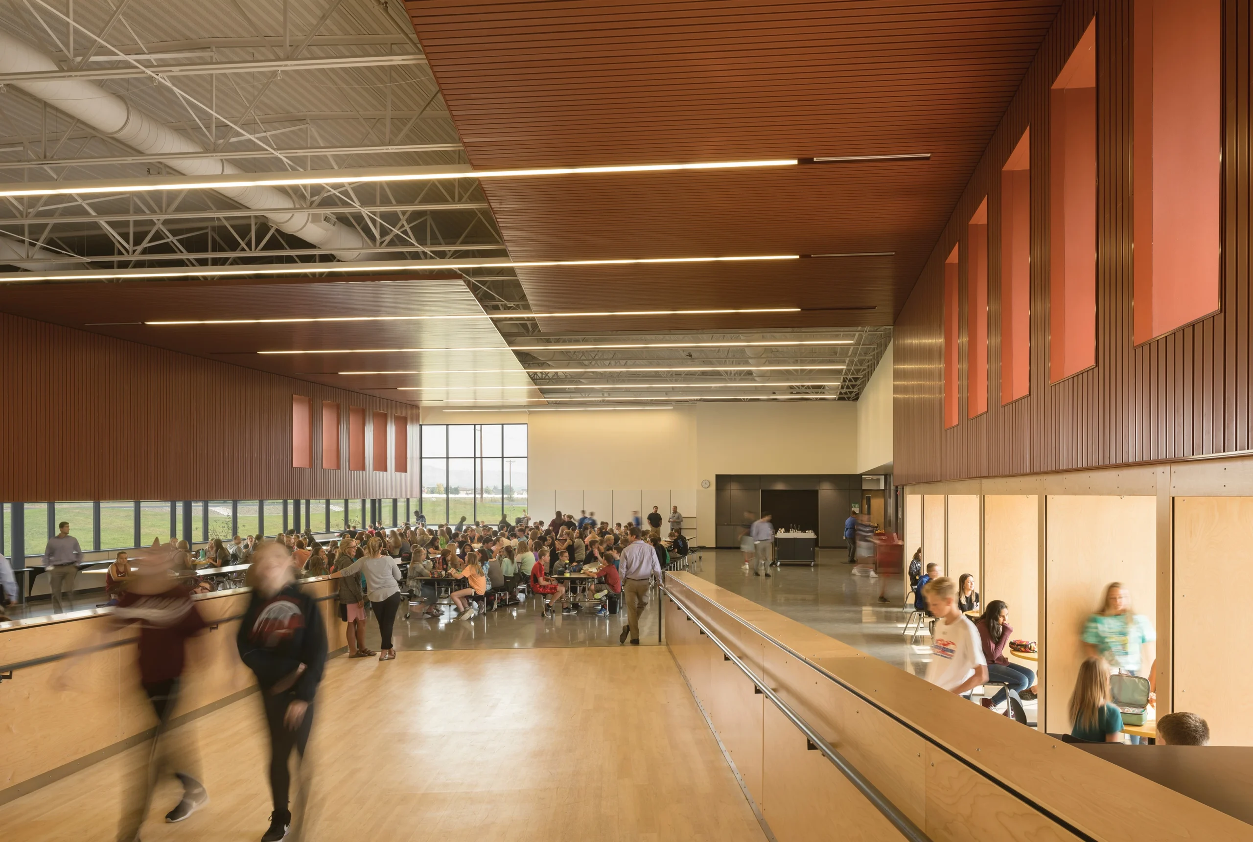 Interior view of Ben Steele Middle School’s bustling commons area, showcasing a vibrant cafeteria filled with students. The wood-accented ceiling and expansive windows provide natural light, while cozy study nooks along the wall offer spaces for quiet reflection.