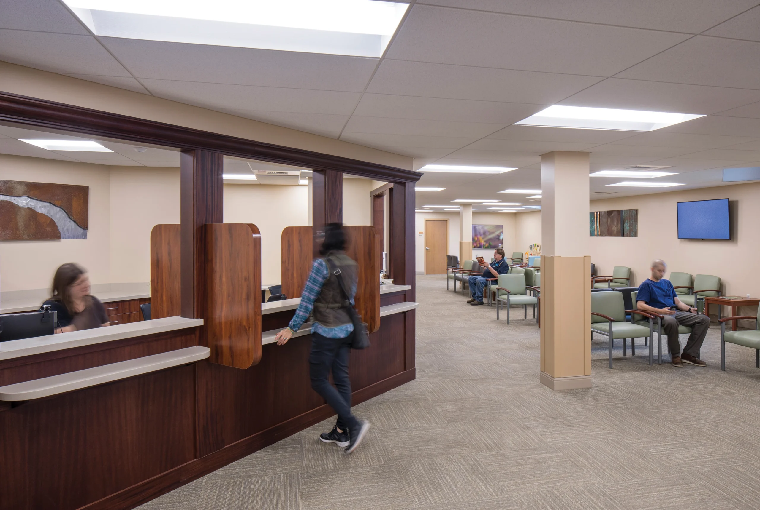 A spacious healthcare waiting area with a reception desk, check-in windows, and ample seating. The neutral color palette and soft lighting create a welcoming and calming environment for patients.