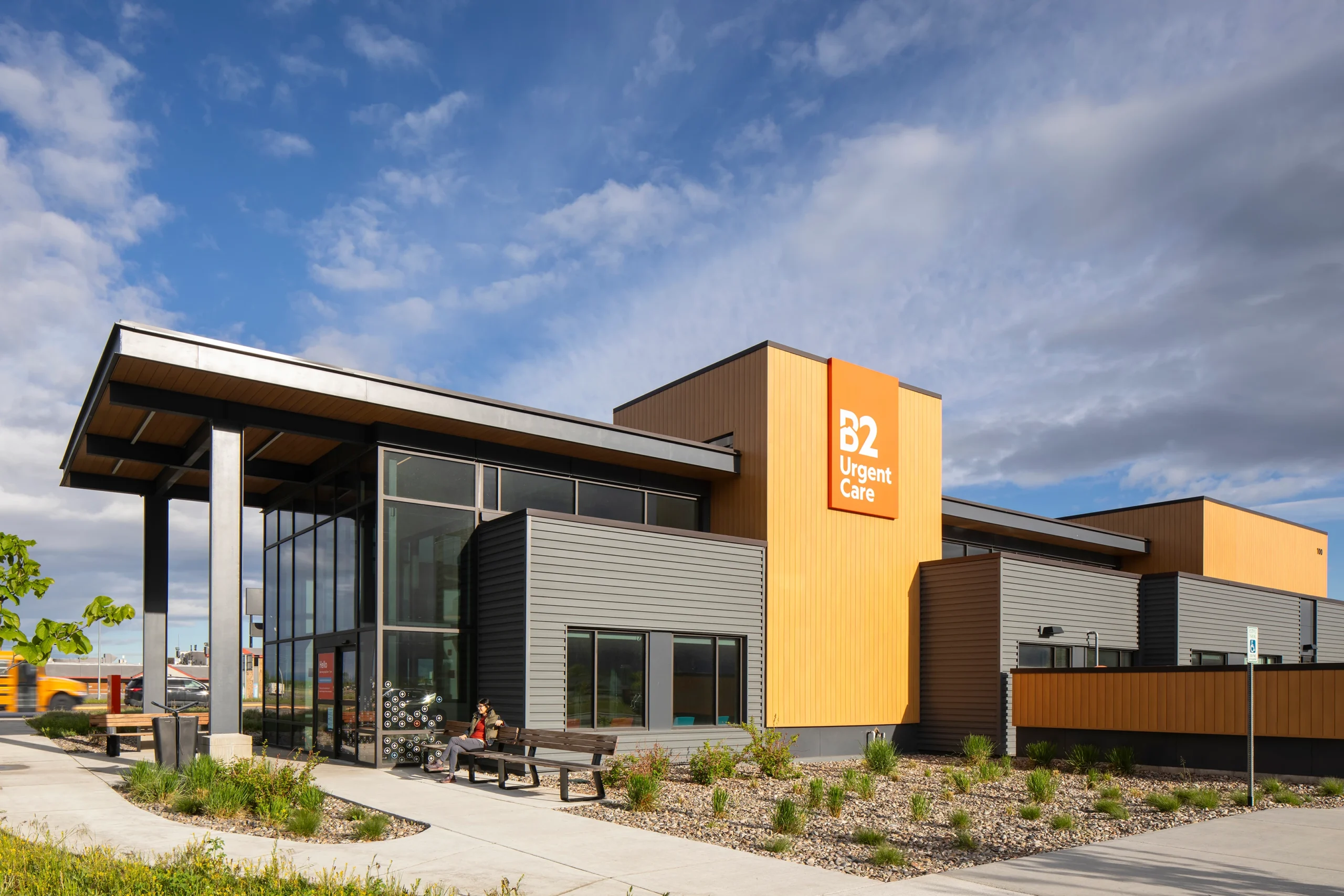 The entrance of B2 Urgent Care features a modern design with warm wood paneling, dark metal siding, and a covered glass entryway, creating an inviting and functional healthcare environment.